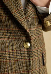 Pruun ruuduline bleiser, mis on valmistatud tekstureeritud kangast, kuldsete nuppudega, millel on emblēm. Ruutude seas on sinise ja burgundia varjundeid.