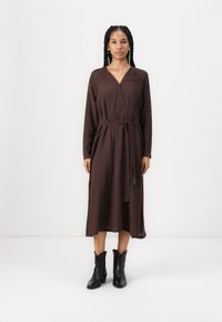 Robe enveloppante marron à manches longues, avec une ceinture nouée à la taille et une silhouette fluide. Associée à des bottines noires. 