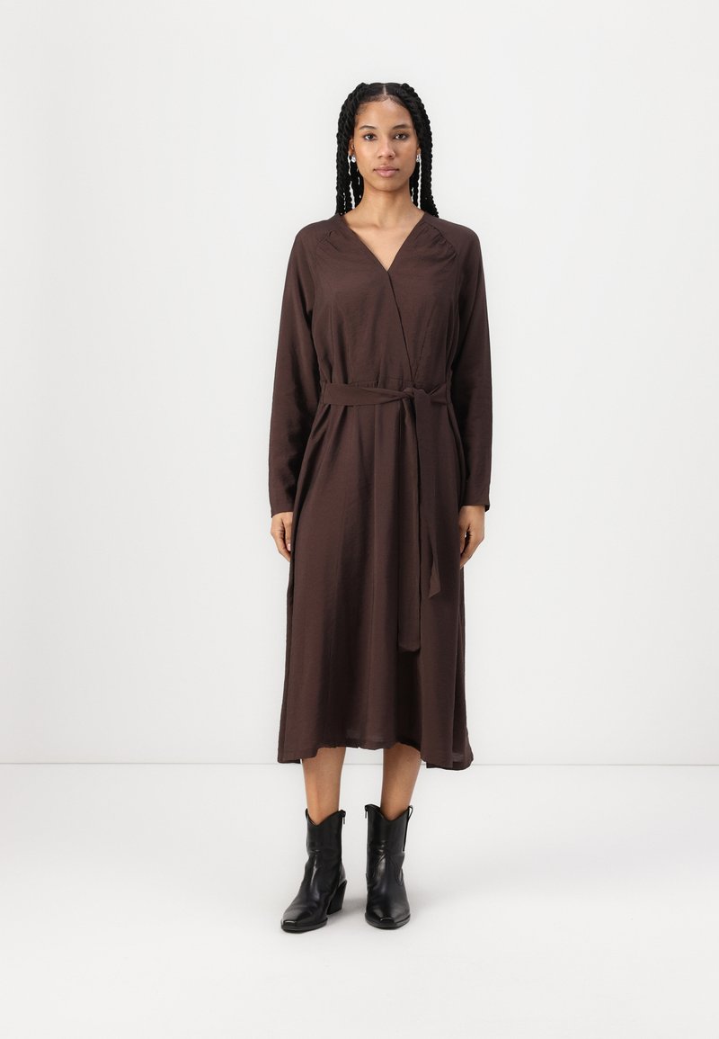Robe enveloppante marron à manches longues, avec une ceinture nouée à la taille et une silhouette fluide. Associée à des bottines noires. 