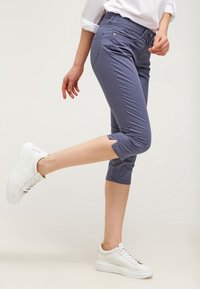 Jasnofioletowe spodnie capri wykonane z gładkiego materiału o dopasowanym kroju. Posiadają mankiety na końcach nogawek i kieszenie z przodu, w połączeniu z białymi sneakersami.