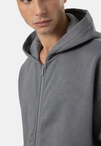 Felpa grigia con zip realizzata in tessuto morbido, con tasca frontale e logo in rilievo sul petto. Cappuccio e dettagli della zip visibili.