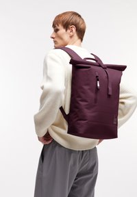 Zaino roll-top color bordeaux con spallacci regolabili, tasca frontale e dettaglio del logo. Realizzato in materiale resistente con una texture liscia.