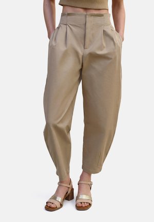Mujer con pantalones beige plisados de cintura alta y pierna estrecha, y sandalias beige de tacón ancho con tiras en el tobillo.