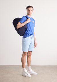 T-shirt en coton bleu avec un logo, shorts bleu clair et baskets blanches. Sac de sport bleu foncé avec une bandoulière. Design minimaliste.