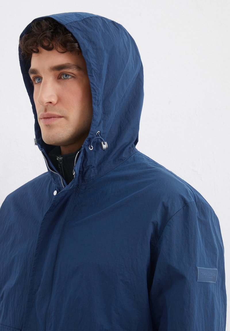 Marineblauwe gevoerde jas van lichtgewicht stof, voorzien van een drukknoopsluiting, trekkoorden op de capuchon en een logopatch op de mouw.