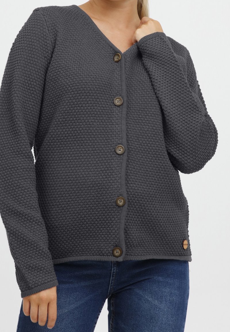 Šedý pletený cardigan s výstřihem do V, texturovaná látka a šest hnědých knoflíků. Na boku malá nášivka s logem. Nošený přes modré džíny.