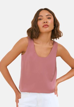 Femme aux cheveux ondulés jusqu'aux épaules porte un haut mauve sans manches et un pantalon blanc, debout, les mains sur les hanches, sur fond blanc.