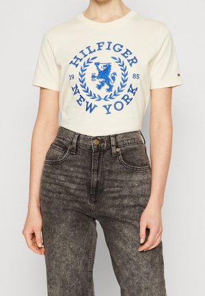 Persona con una camiseta crema con la inscripción azul "Hilfiger New York 1985" y un escudo con un león, combinada con jeans negros desteñidos de tiro alto.