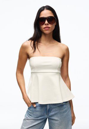 Kobieta w dużych ciemnych okularach przeciwsłonecznych, białym, bezramkowym topie typu peplum i niebieskich dżinsach, stojąca z jedną ręką w kieszeni na białym tle.