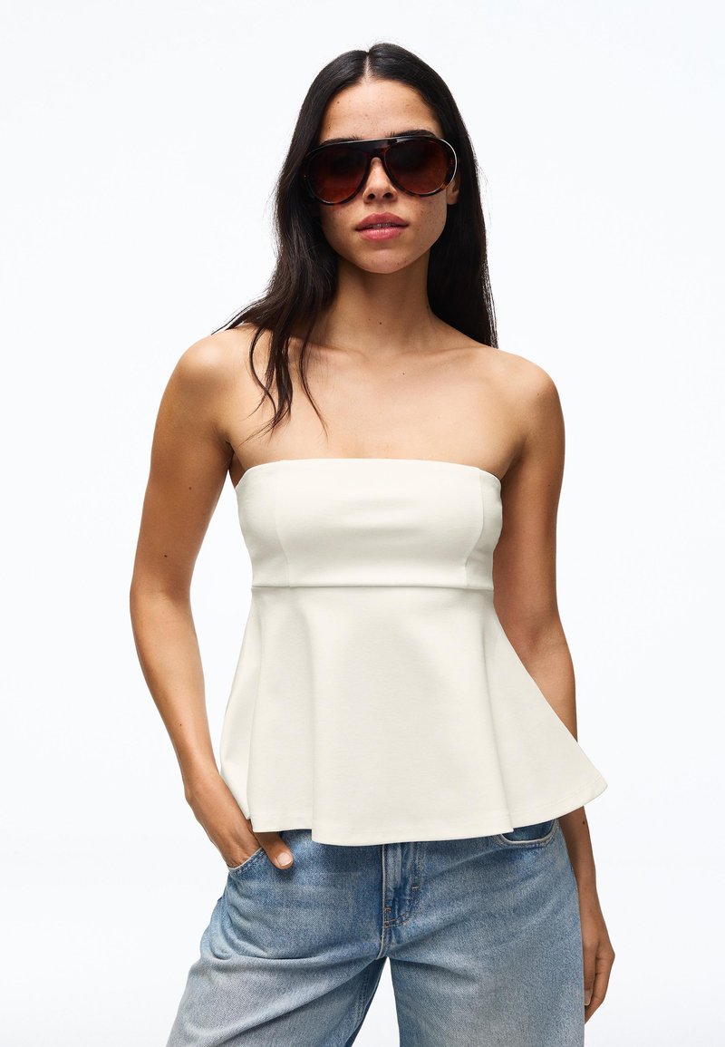 Femme portant de grandes lunettes de soleil foncées, un top blanc bustier à basque et un jean bleu, debout avec une main dans la poche sur fond blanc.