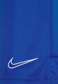 Tissu bleu avec un logo Nike swoosh blanc cousu près du bord inférieur sur le panneau latéral d'un vêtement.