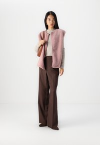 Vesta roz din fleece cu deschidere frontală, asociată cu un pulover ușor și pantaloni maro cu picioare largi. Modelul ține o mică geantă de umăr maro.