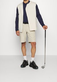 Man i beige shorts, vit väst, marinblå långärmad skjorta, svarta skor, hållande en golfklubba stående mot en enkel bakgrund.