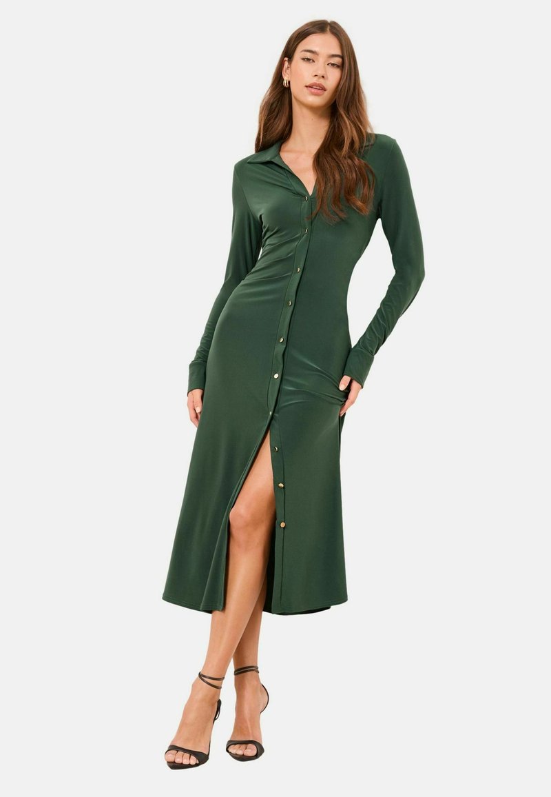 Rochie verde cu mâneci lungi și nasturi, realizată dintr-un material neted; are o fustă cu despicătură până la coapsă și nasturi aurii în față.