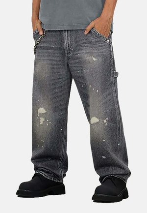 Jeans grigi in denim larghi e consumati con schizzi di vernice, orlo sfrangiato, accessori metallici e una catena decorativa sulla tasca.