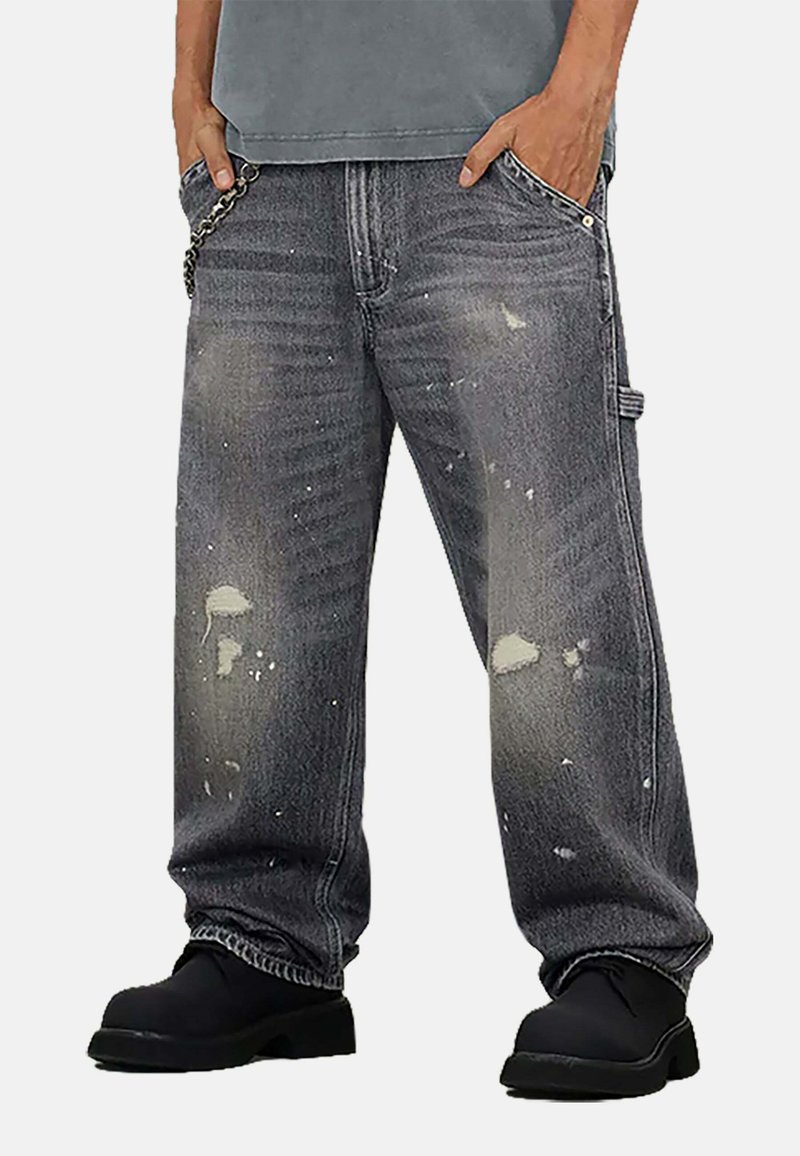 Jeans grigi in denim larghi e consumati con schizzi di vernice, orlo sfrangiato, accessori metallici e una catena decorativa sulla tasca.