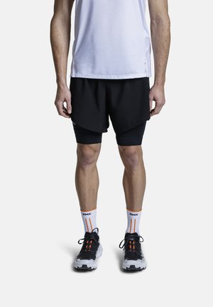 Man die zwarte gelaagde hardloopshorts draagt, witte sportsokken met oranje strepen, zwarte hardloopschoenen en een wit hemd, rechtopstaand.