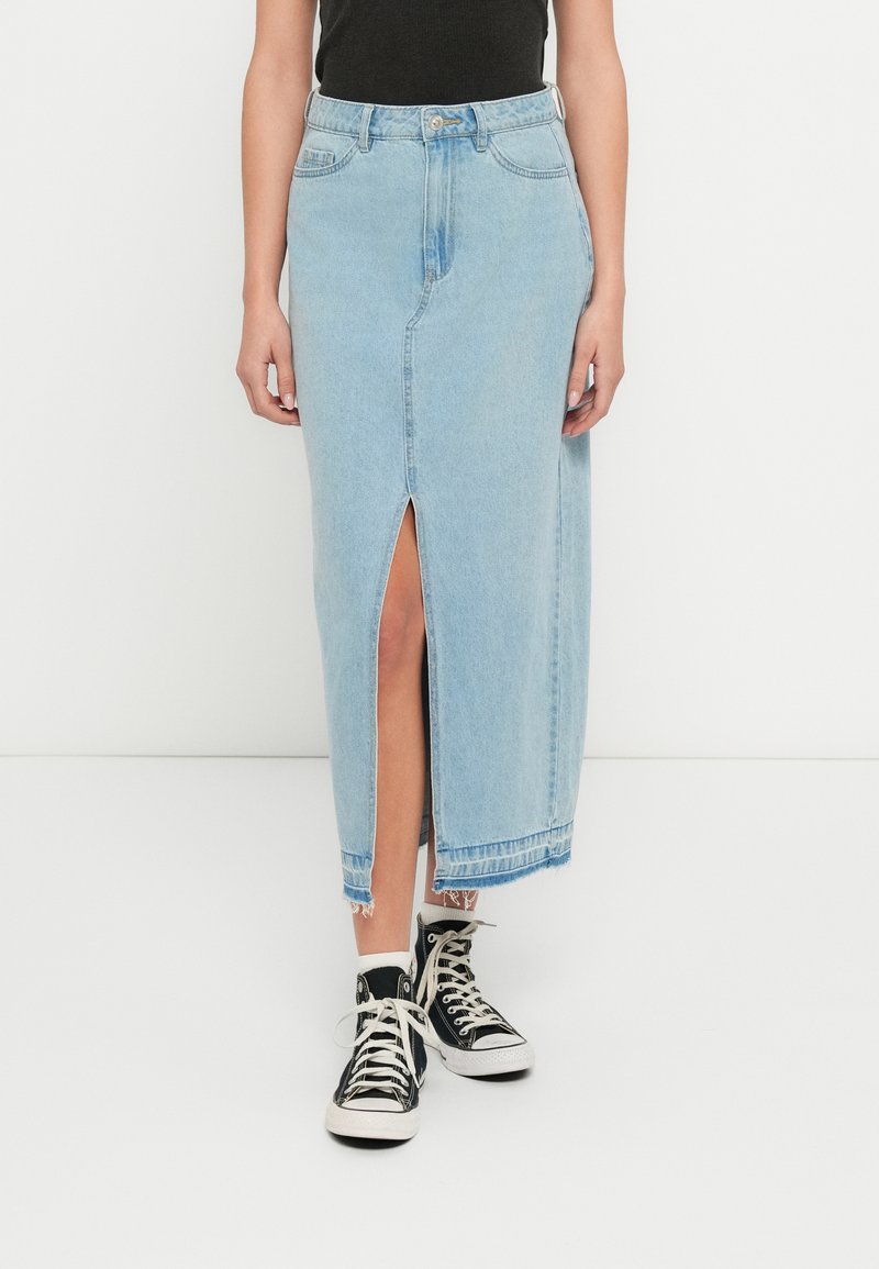 Lichtblauwe denim midi-rok met een voorste split, afgewerkte zoom en een lichte rafeling. Draagt zwarte hoge sneakers en een nauwsluitende zwarte top.