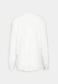 Chemise blanche à manches longues avec poignets boutonnés, vue de dos sur un fond clair uni.