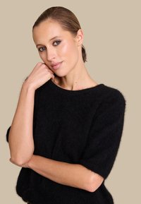 Svart, loose-fitting strikkegenser med korte ermer, laget av mykt, strukturert stoff. Modellen poserer med hånden nær haken mot en beige bakgrunn.