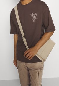 Muž v hnědém grafickém tričku a béžových cargo kalhotách, držící béžovou křížovou tašku s nastavitelným popruhem.