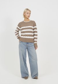 Brązowy i biały sweter w paski, dziergany, z fakturowanym wzorem diamentów, w połączeniu z jasnoniebieskimi spodniami w szerokim kroju i beżowymi butami.