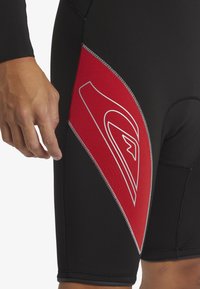 Kort våtdräkt med svart bas, röd sidopanel med vita detaljer, flatlock-sömmar och en åtsittande design för vattenaktiviteter.