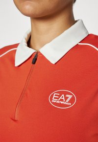 Raudona polo marškinėliai, pagaminti iš lygios medžiagos, su balta apykakle, priekiniais užtrauktukais ir išsiuvinėtu EA7 Emporio Armani logotipu.