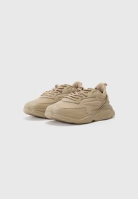 Beige sneakers feature a textured fabric upper, chunky rubber sole, rounded toe, and lace-up design with a loop at the heel for easy wear.