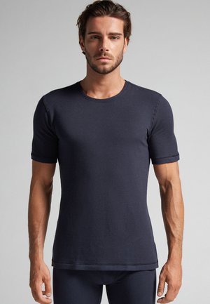 Man met kort bruin haar en baard, die een aansluitend donkerblauw T-shirt met korte mouwen en bijpassende broek draagt tegen een effen achtergrond.