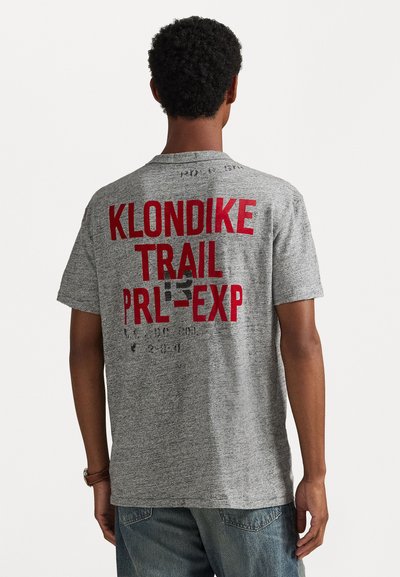 Harmaa lyhythihainen t-paita, jonka takana on rohkeaa punaista tekstiä, joka lukee "KLONDIKE TRAIL PRL-EXP." Kangas on pehmeä ja tekstuuri on miellyttävä.