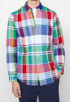 Homme portant une chemise à manches longues avec un grand motif à carreaux multicolore et un pantalon bleu foncé, debout avec une main dans la poche.