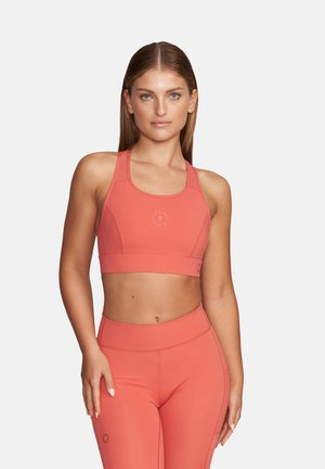 Kobieta o prostych włosach, ubrana w pasujący koralowy sportowy biustonosz i legginsy z wysokim stanem, stojąca na jednolitym tle.