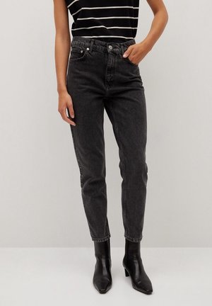 Person iført sorte high-waist jeans, sort stribet ærmeløs top og sorte ankelstøvler med hæl, stående mod en ensartet lys baggrund.