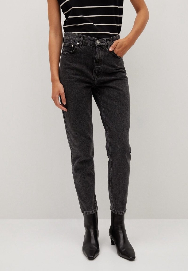 Personne portant un jean taille haute noir, un top sans manches rayé noir et des bottines noires à talons, debout devant un fond clair uni.