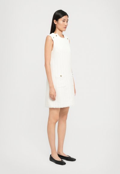 Biała sukienka bez rękawów z teksturowanej tkaniny, z dwiema przednimi kieszeniami, złotymi guzikami i postrzepionym dołem. Noszona z czarnymi płaskimi butami.