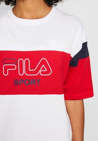 Camiseta deportiva FILA con bloques de color rojo y blanco en horizontal, cuello redondo con canalé y texto de logo en colores contrastantes.