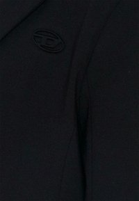 Tissu noir, texture lisse, coupe ajustée. Présente un logo ovale brodé en haut à gauche. Aucune motif ni accent supplémentaire visible.