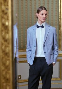 Gaiši zils, teksturēts žakets ar diviem podziņām pār balto kreklu; melnas oficiālas bikses. Tašs kaklasaites akcentē oficiālo apģērbu.