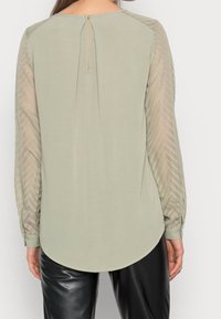 Blusa verde chiaro a maniche lunghe con un motivo a zigzag trasparente e testurizzato sulle maniche, scollatura rotonda e dettaglio a chiusura con bottone sul retro.