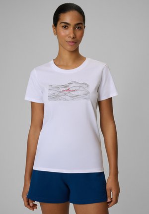 Kobieta o ciemnych włosach w białym, graficznym t-shircie z napisem „STEP INTO THE WILD” oraz granatowych szortach stoi na szarym tle.