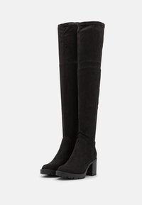 Botas de ante negras por encima de la rodilla con tacones altos gruesos y suelas de goma estriadas, de pie una al lado de la otra sobre un fondo blanco.