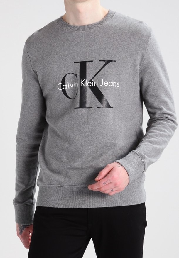 Personne portant un sweat-shirt gris Calvin Klein Jeans avec un pantalon noir, main montrant de petits tatouages sur les doigts.