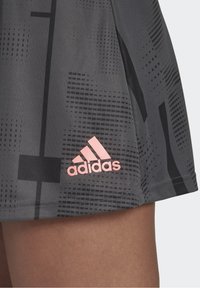 Grå atletisk kjol med ett geometriskt mönster i svart nyanser, accentuerad av en rosa Adidas-logotyp nära fållen. Tillverkad av lättviktsmaterial.
