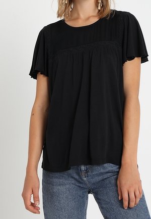 Frau trägt eine schwarze Bluse mit kurzen Ärmeln und Spitzendetails, kombiniert mit blauen Jeans, gezeigt von den Schultern bis zur Hüfte.