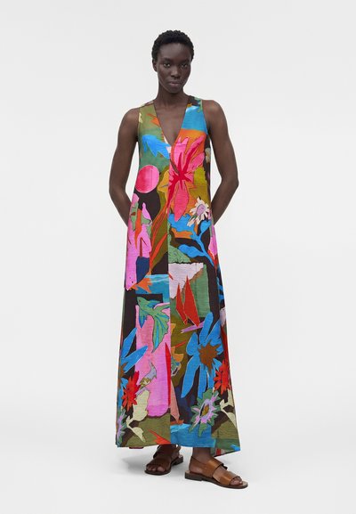 Γυναίκα που φοράει ένα αμάνικο, πολύχρωμο φόρεμα μέχρι το πάτωμα με βαθύ V-λαιμόκοψη και αφηρημένα ανθισμένα σχέδια, σε συνδυασμό με καφέ σανδάλια ανοιχτών δακτύλων.