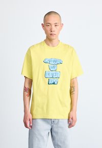 Kollane puuvillane T-särk, millel on sinine graafiline trükk tekstiga "Carhartt WIP Detroit USA." Lõdva lõikega ja lühikeste käistega.