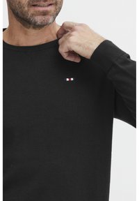 Juodas megztinis su mėginiu apykakle, lygia tekstūra ir maža siuvinėta logotipu raudona, balta ir mėlyna spalvomis ant krūtinės.