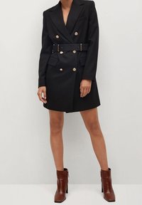 Schwarzes, doppelt-knopfbares Blazer-Kleid mit goldenen Knöpfen, breiten Revers und passendem Gürtel, ausgestattet mit seitlichen Taschen und taillierter Passform. Braune Stiefeletten.
