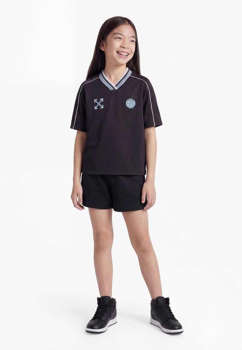 Νεαρή κοπέλα που χαμογελά, φοράει μαύρη αθλητική φόρμα με λογότυπα, μαύρο σορτς και μαύρα αθλητικά παπούτσια, στέκεται μπροστά σε λευκό φόντο.